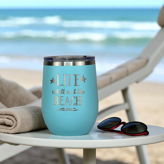 metal teal wine tumbler on a side tale with sunglasses next to a beach lounge chair tumbler.  Soft breeze vibe with ocean blur in background.