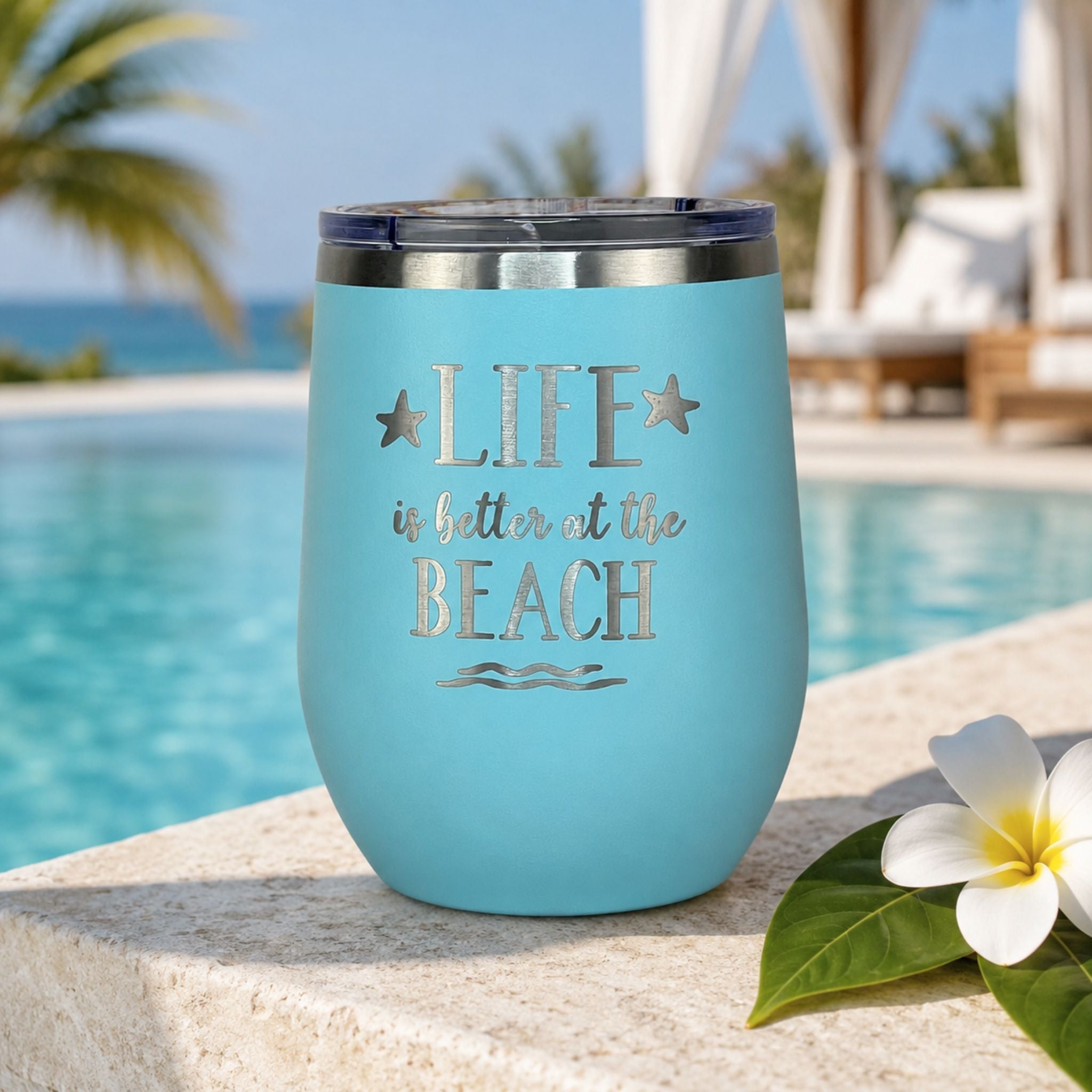 teal metal tumbler with 'Life is better at the Beach' text by a poolside with palm trees and flowers.
