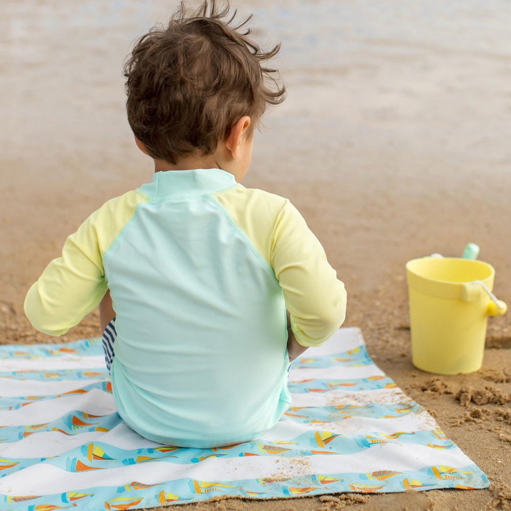 little boy on a sandy beach sitting on a light blue striped with sailboat pattern, quick dry, sand-free kids beach towel