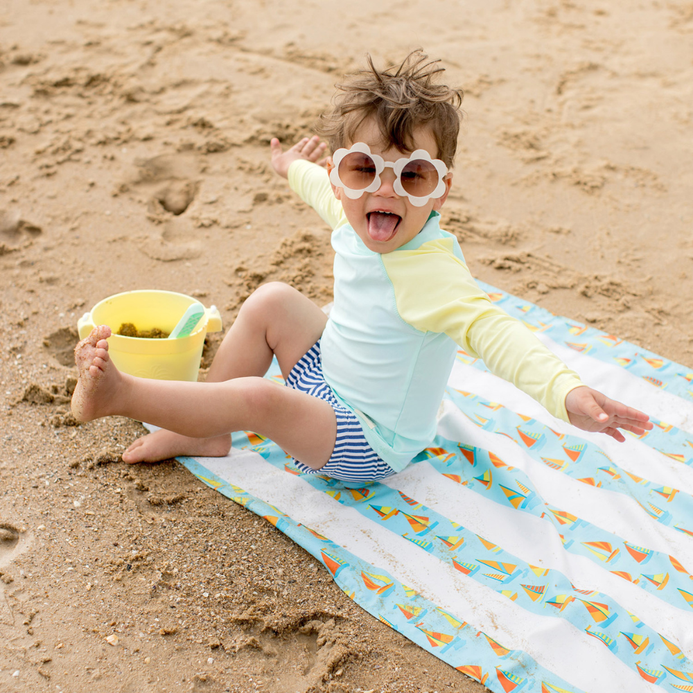 adorable boy on sandy beach with bucket, sitting on light blue striped with sailboat pattern, quick dry, sand-free kids beach towel laughing