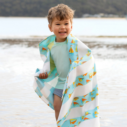 little boy smiling standing in front of water on a sandy beach wrapped in light blue striped with sailboat pattern, quick dry, sand-free kids beach towel