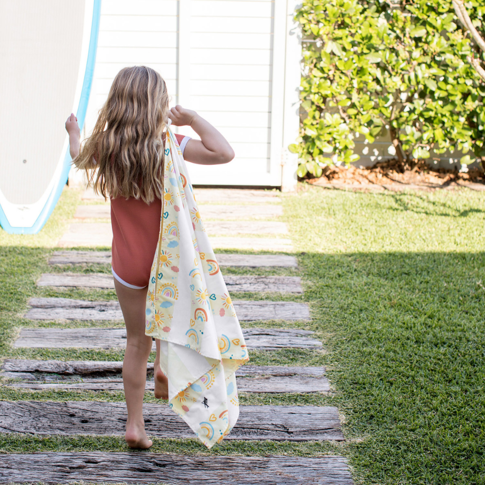 girl walking on wood pathway holding pastel, yellow striped rainbow pattern quick dry sand free kids beach towel 