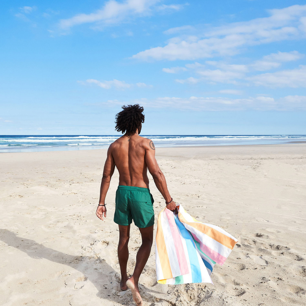 man on sandy beach holding bright, pastel rainbow striped quick dry, sand free beach towel