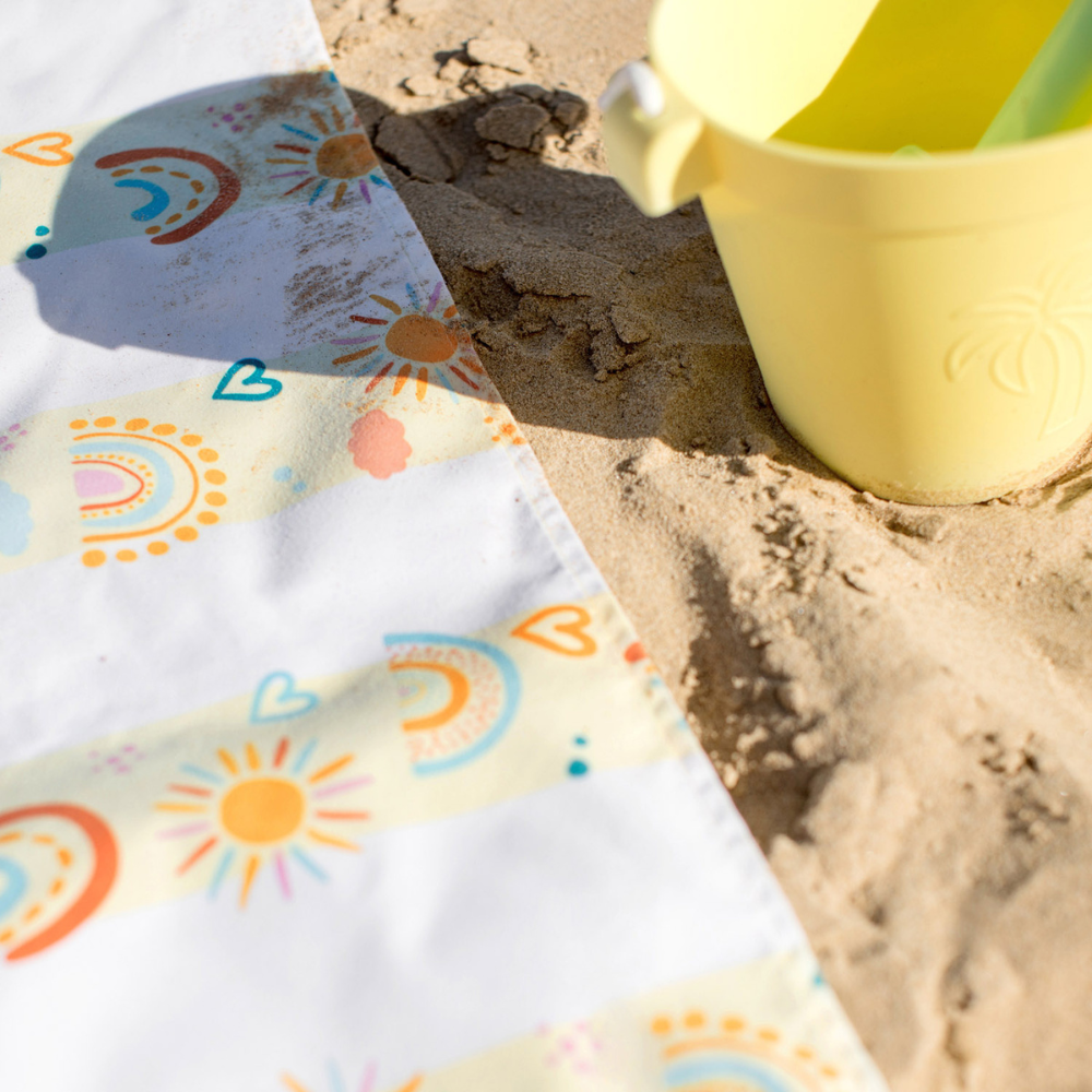 pastel, yellow striped rainbow pattern quick dry sand free kids beach towel on a sandy beach with bucket