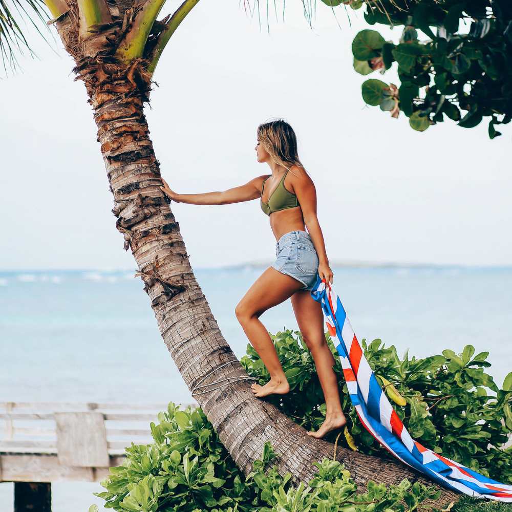 woman standing on palm tree holding patriotic, red, white and blue quick dry and sand free beach towel